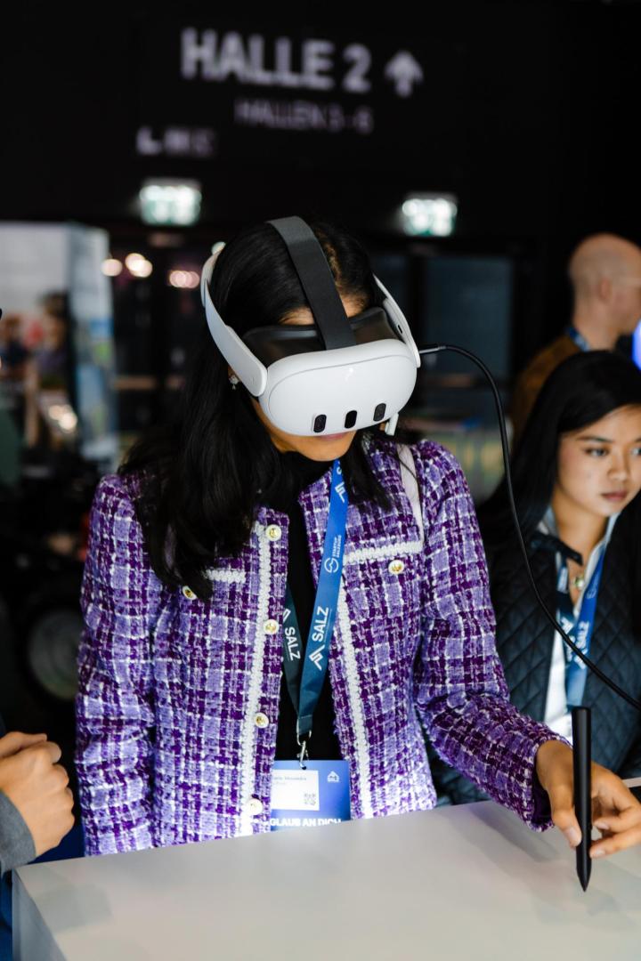 Eine Frau trägt eine weiße VR-Brille, steht vor einem Tisch und schreibt mit einem schwarzen Stift auf die Oberfläche.  Sie trägt einen lila Blazer im Channel-Stil.