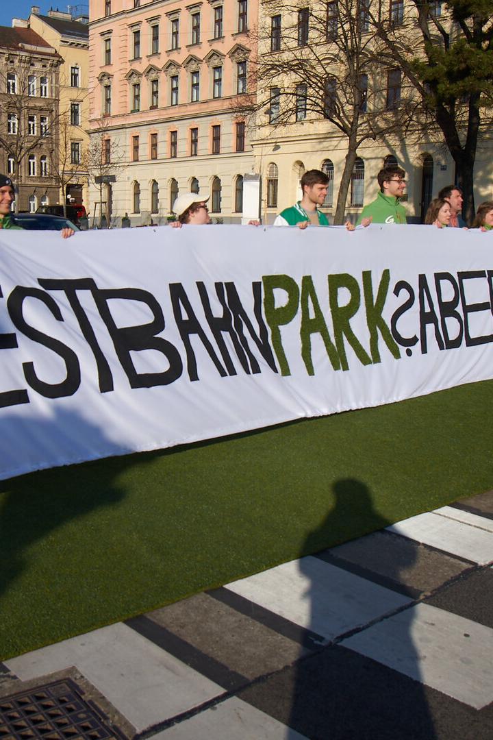 Mehrere Menschen halten ein langes Transparent mit der Aufschrift „WESTBAHNPARK? ABER RICHTIG!“ auf einer Straße.