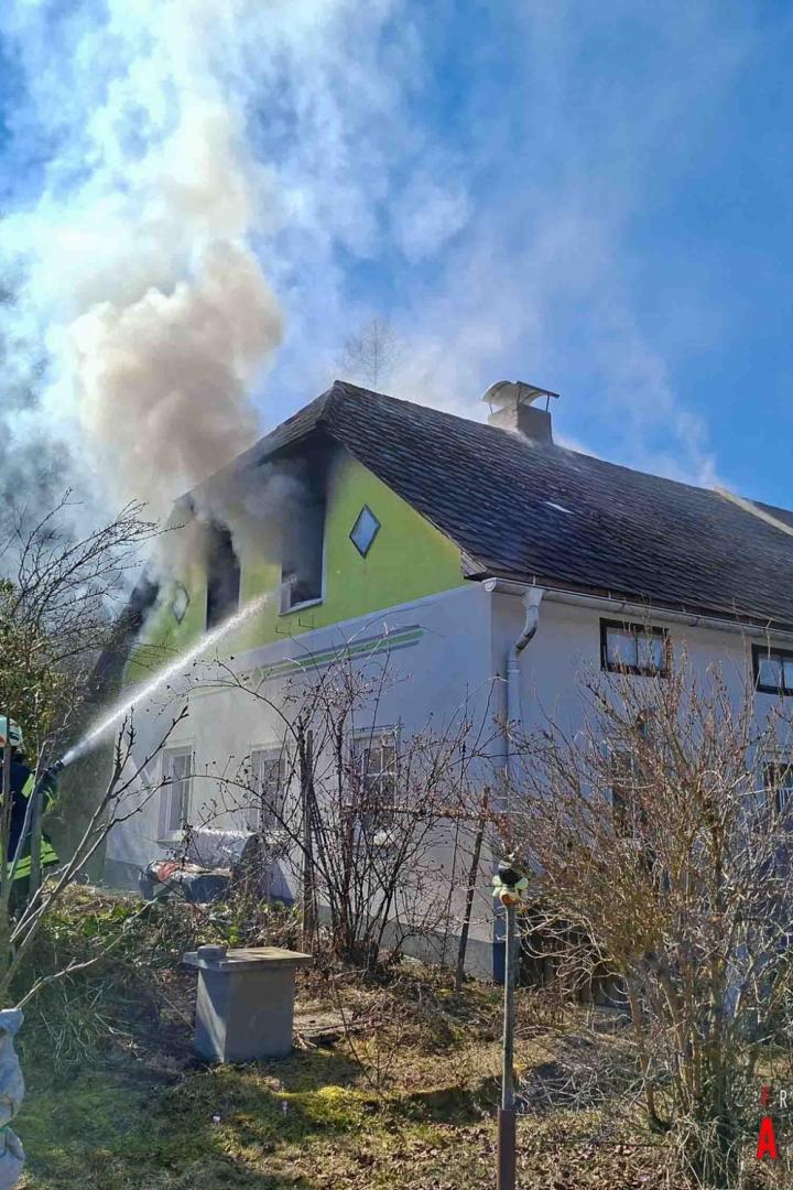 Zwei Feuerwehrmänner halten einen Schlauch und richten den Strahl auf das Fenster eines Wohnhauses, aus dem Rauch aufsteigt.
