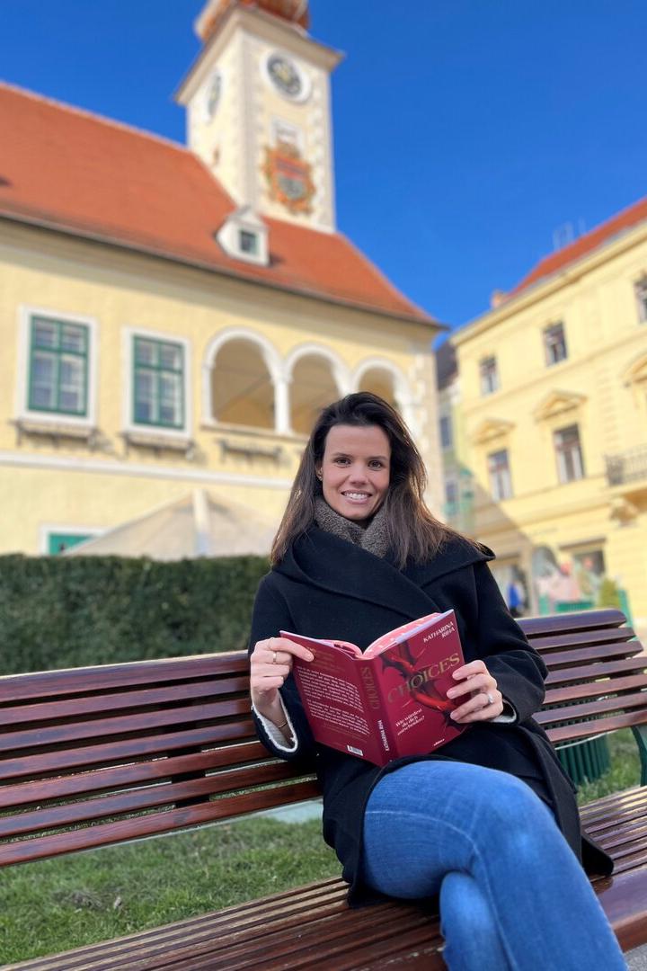 Autorin Katharina Riha sitzt mit ihrem aufgeschlagenen Buch in den Händen auf einer Parkbank.