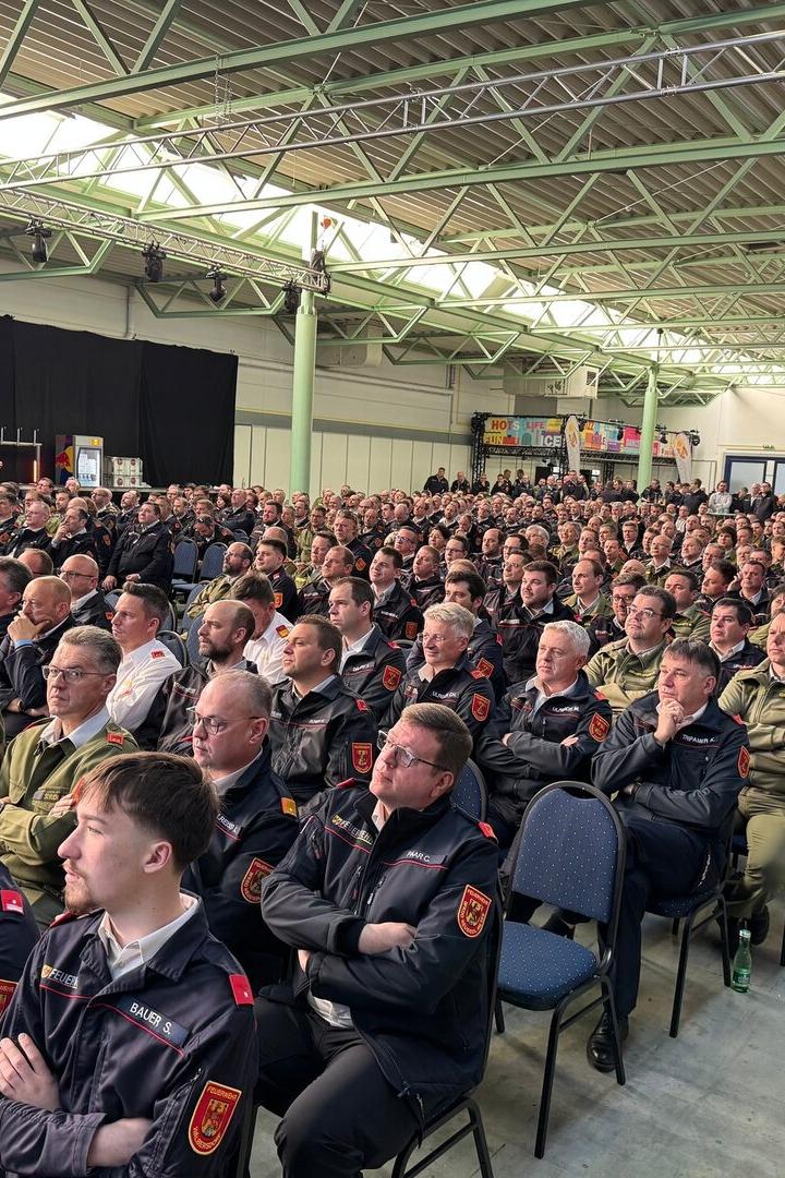 Viele Feuerwehrleute in Uniform sitzen dicht gedrängt in einer großen Halle und hören aufmerksam zu.