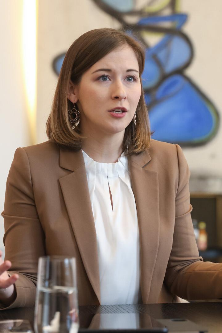 Eine Frau mit braunen Haaren und beigem Blazer spricht gestikulierend an einem Tisch in einem modernen Büro.