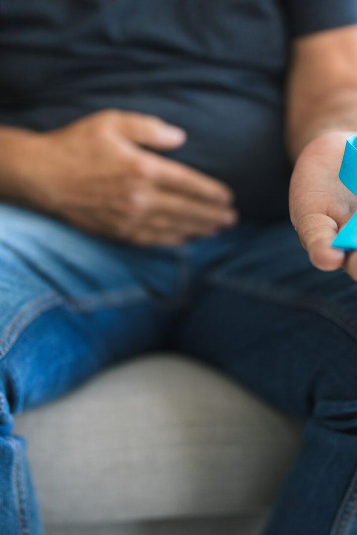 Ein Mann sitzt auf einem Sofa, hält eine hellblaue Schleife in der Hand und fasst sich an den Bauch.