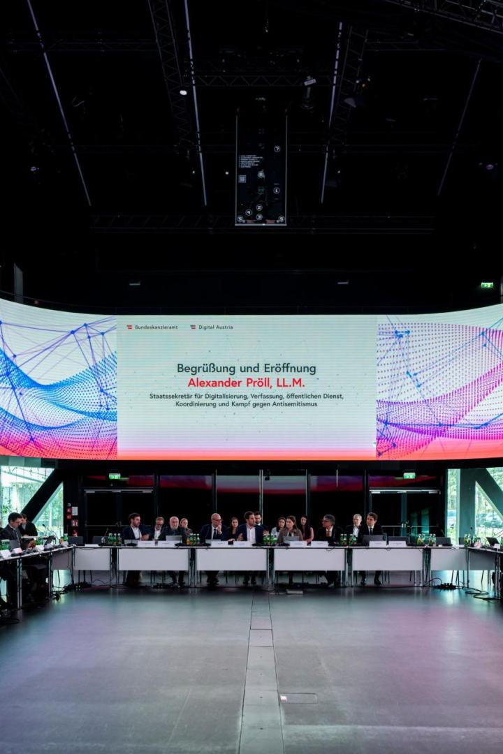 Viele Menschen sitzen an einem großen runden Konferenztisch, über ihnen eine große digitale Anzeigetafel mit buntem Muster.