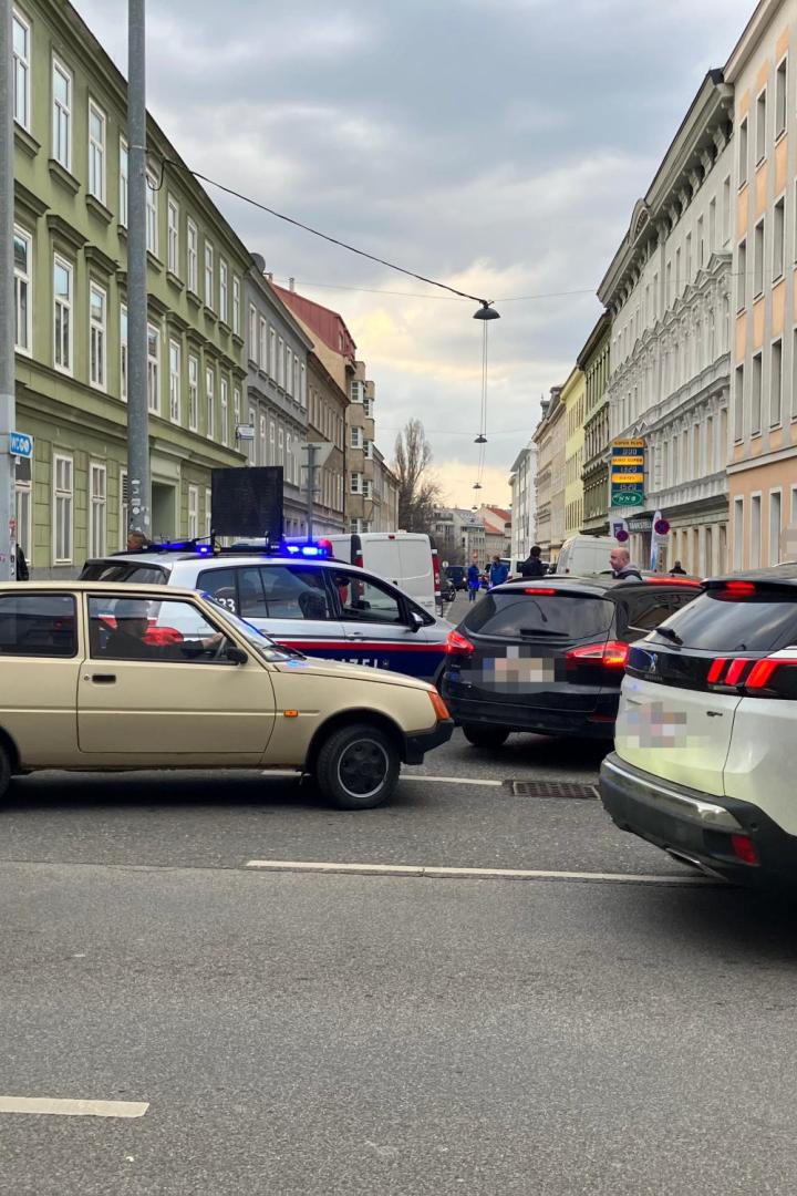 Mehrere Autos und Polizisten stehen an einer Straßensperre in einer Stadt mit Wohnhäusern und einem FPÖ-Stand.