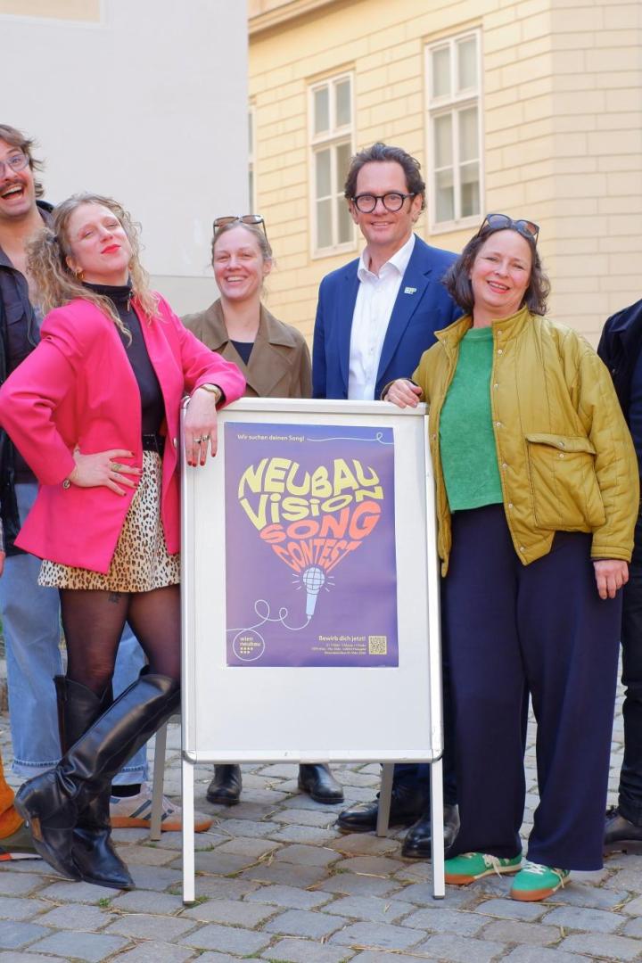 Mehrere bunt gekleidete Menschen, darunter Neubau Bezirksvorsteher Reiter, stehen vor einem Plakatschild auf Pflastersteinen