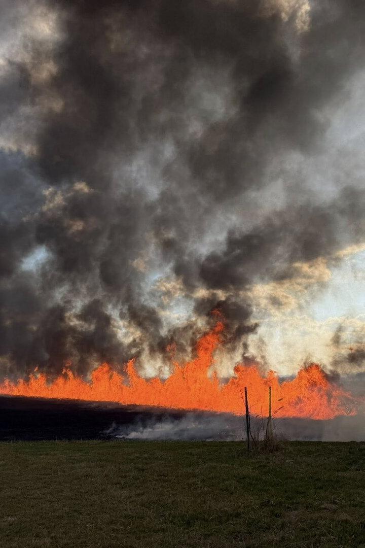 Großes Feuer mit dichten schwarzen Rauchwolken breitet sich auf einem Feld aus, während die Sonne am Himmel steht.