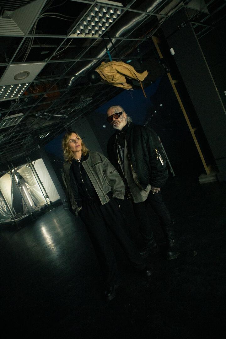 Zwei Personen stehen in einem dunklen Raum mit offenem Deckengebälk, über ihnen hängt eine Schaufensterpuppe kopfüber.