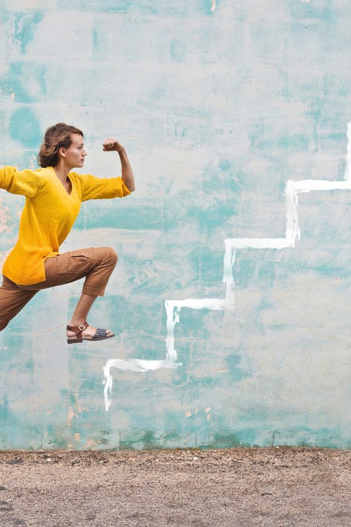 Eine Frau springt dynamisch vor einer Wand mit aufgemalten weißen Treppenstufen nach oben.