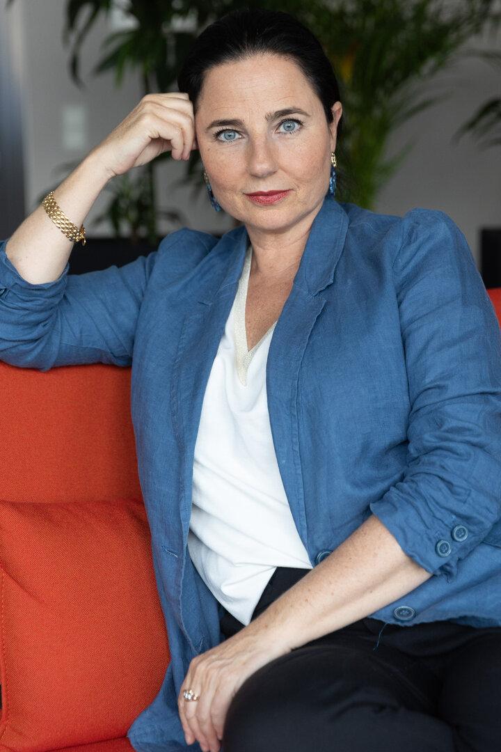 Frau mit dunklen Haaren und blauen Augen sitzt auf einem roten Sofa, trägt einen blauen Blazer und schaut direkt nach vorn.