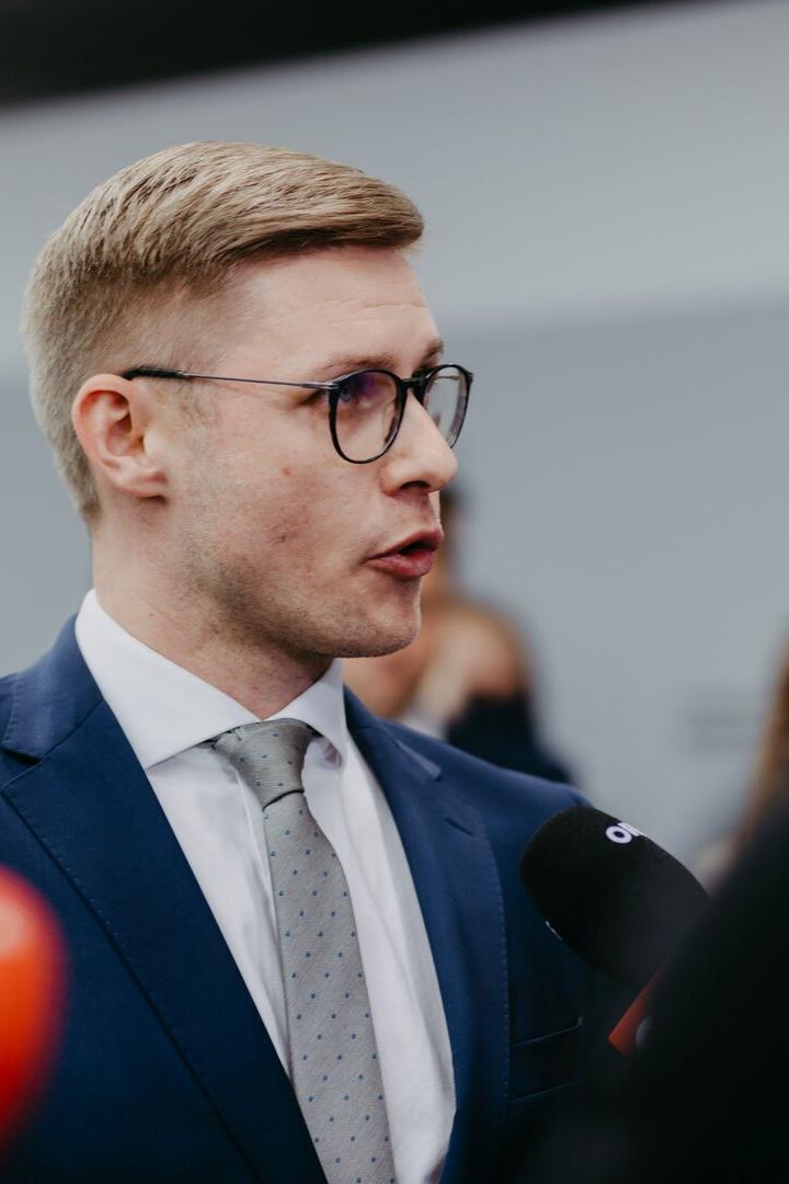 Ein Mann mit Brille und Anzug spricht in Mikrofone vor unscharfem Hintergrund.