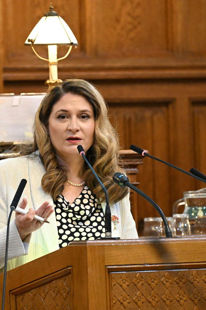 Eine Frau mit Blazer spricht an einem Rednerpult mit Mikrofonen vor holzvertäfeltem Hintergrund.