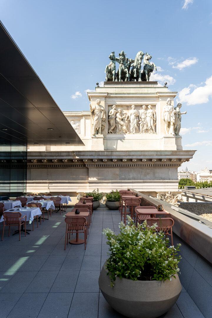 Ein Dachterrassen-Restaurant mit Tischen und Stühlen befindet sich unterhalb eines Gebäudes mit Skulpturen.
