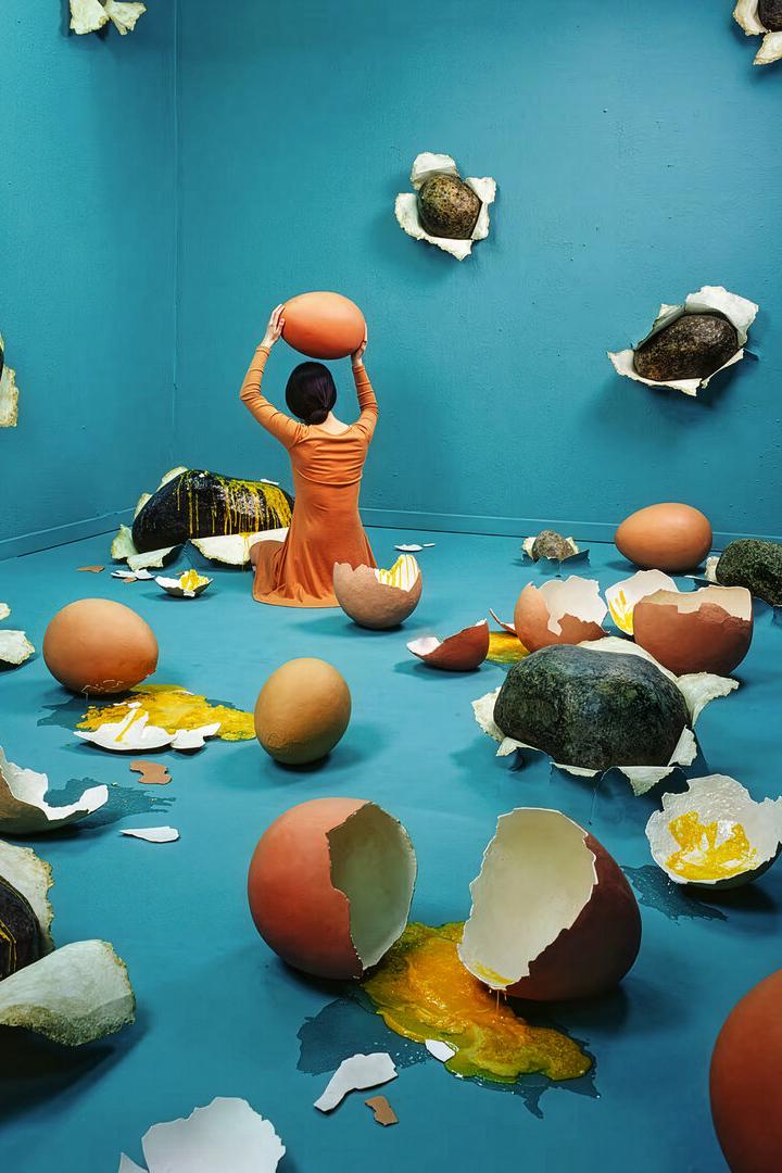 Eine Person in orange sitzt in einem blauen Raum voller zerbrochener Rieseneier und Steine, die aus den Wänden ragen.