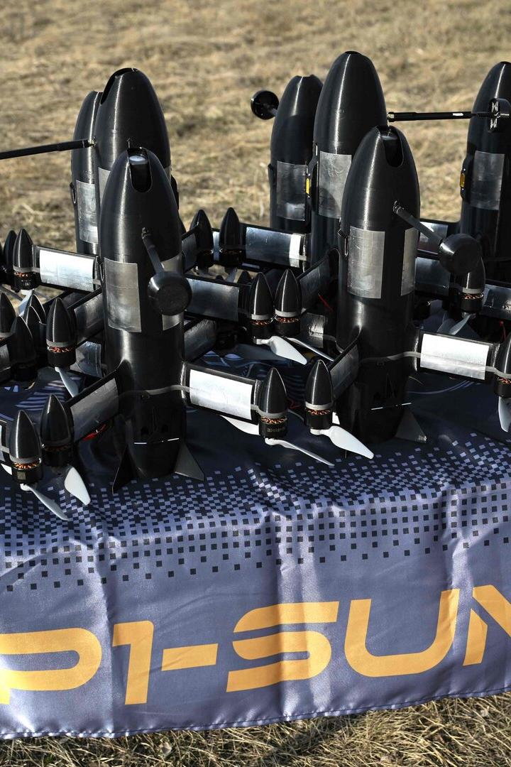 Mehrere schwarze Drohnen mit Propellern stehen auf einem Tisch im Freien auf einer blauen Decke mit gelber Aufschrift.