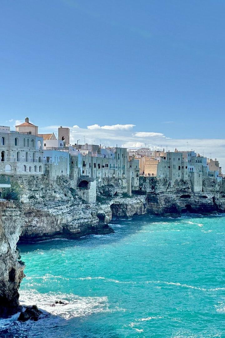 Weiße Häuser thronen auf Klippen über dem türkisfarbenen Meer unter blauem Himmel.
