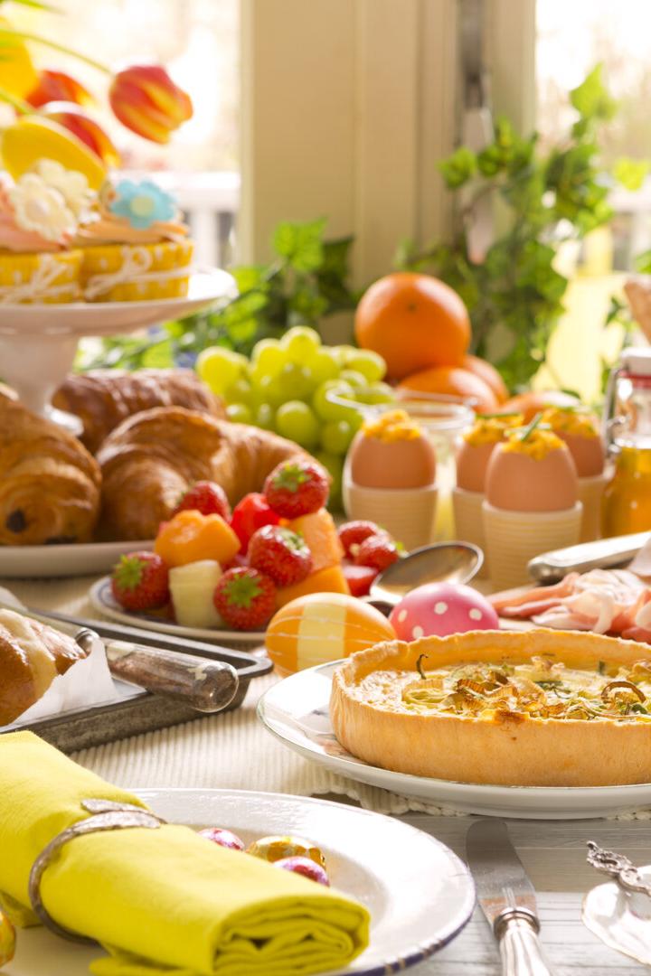 Ein reichhaltiger Brunch mit Gebäck, Quiche, Obst, Eiern und Tulpen steht auf einem Tisch.