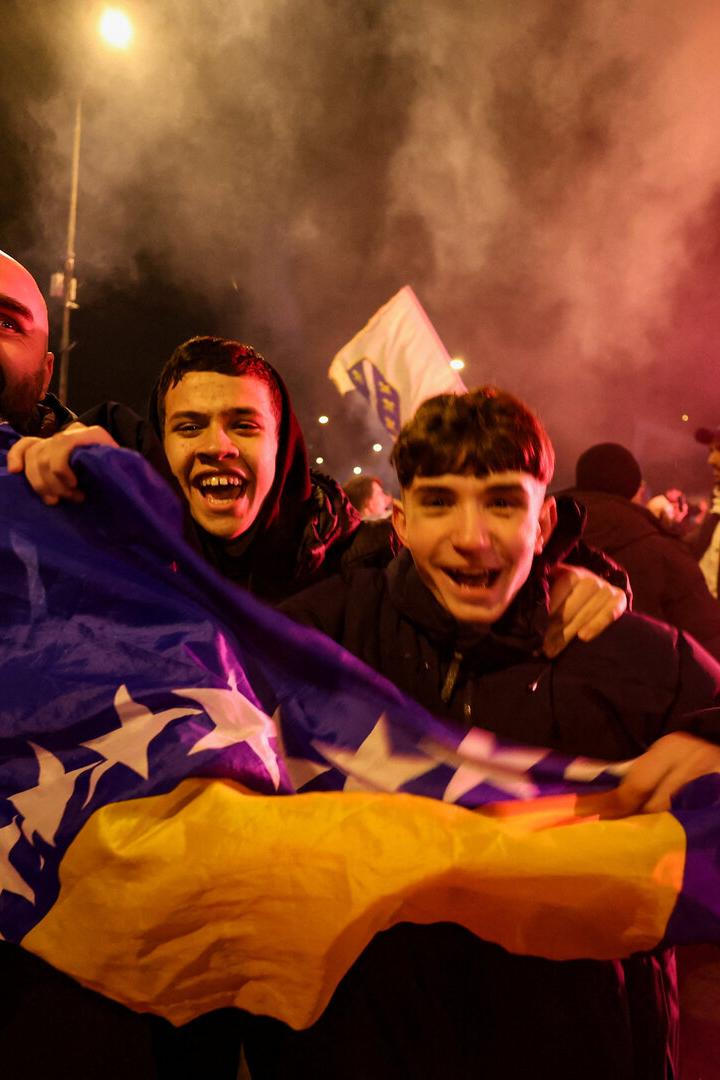 Drei jubelnde Menschen halten eine bosnische Flagge, umgeben von einer feiernden Menschenmenge bei Nacht und Rauch.