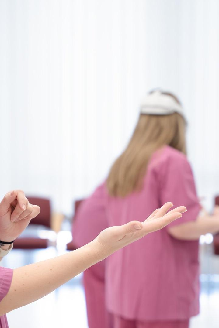 Zwei Personen in rosa Kitteln tragen Virtual-Reality-Headsets und führen gestikulierende Bewegungen in einem hellen Raum aus.