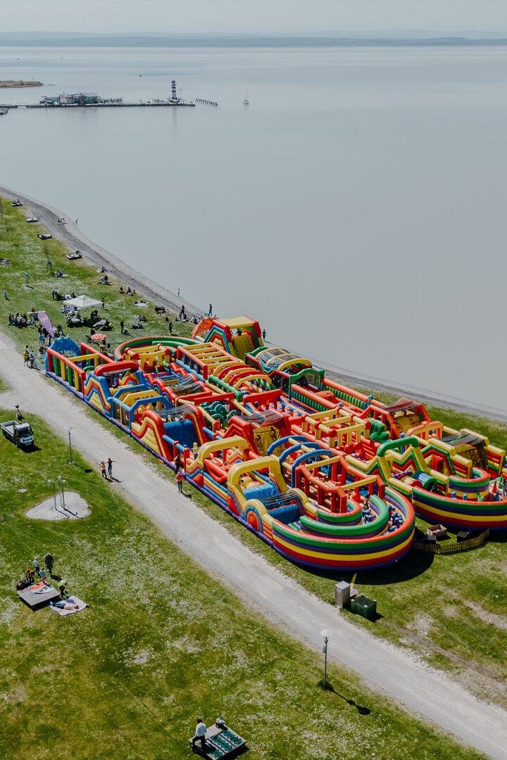 Großer bunter Hüpfburgen-Parcours am Ufer eines Sees, umgeben von Wiese, Wegen und Bäumen mit Menschen.