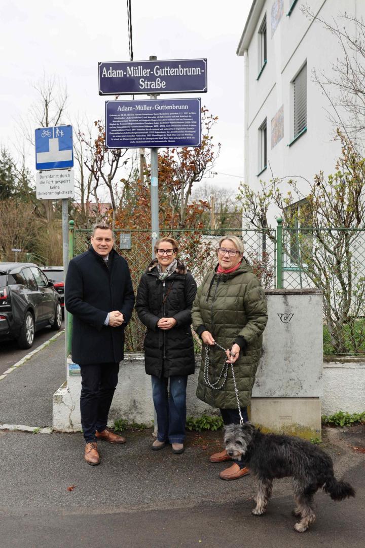 Drei Personen und ein Hund stehen in der Adam-Müller-Guttenbrunn-Straße vor einem Straßenschild mit Informationstafel.
