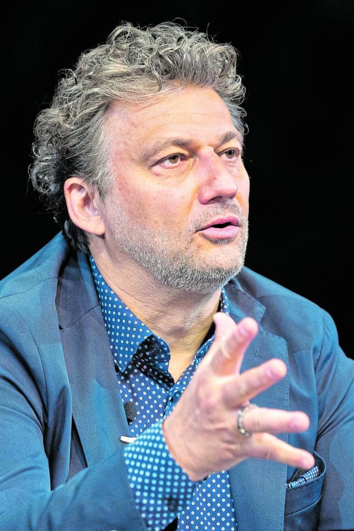 Ein Mann mit lockigen Haaren und Bart gestikuliert mit beiden Händen und trägt ein blaues Sakko über einem gemusterten Hemd.