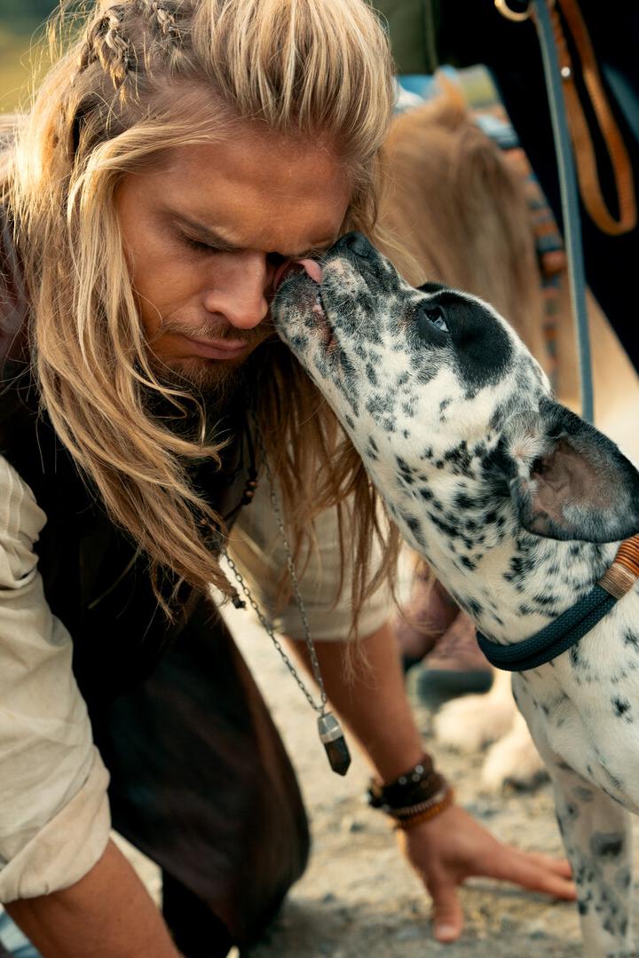 Ein Mann mit langen blonden Haaren wird von einem schwarz-weiß gefleckten Hund im Gesicht abgeschleckt.