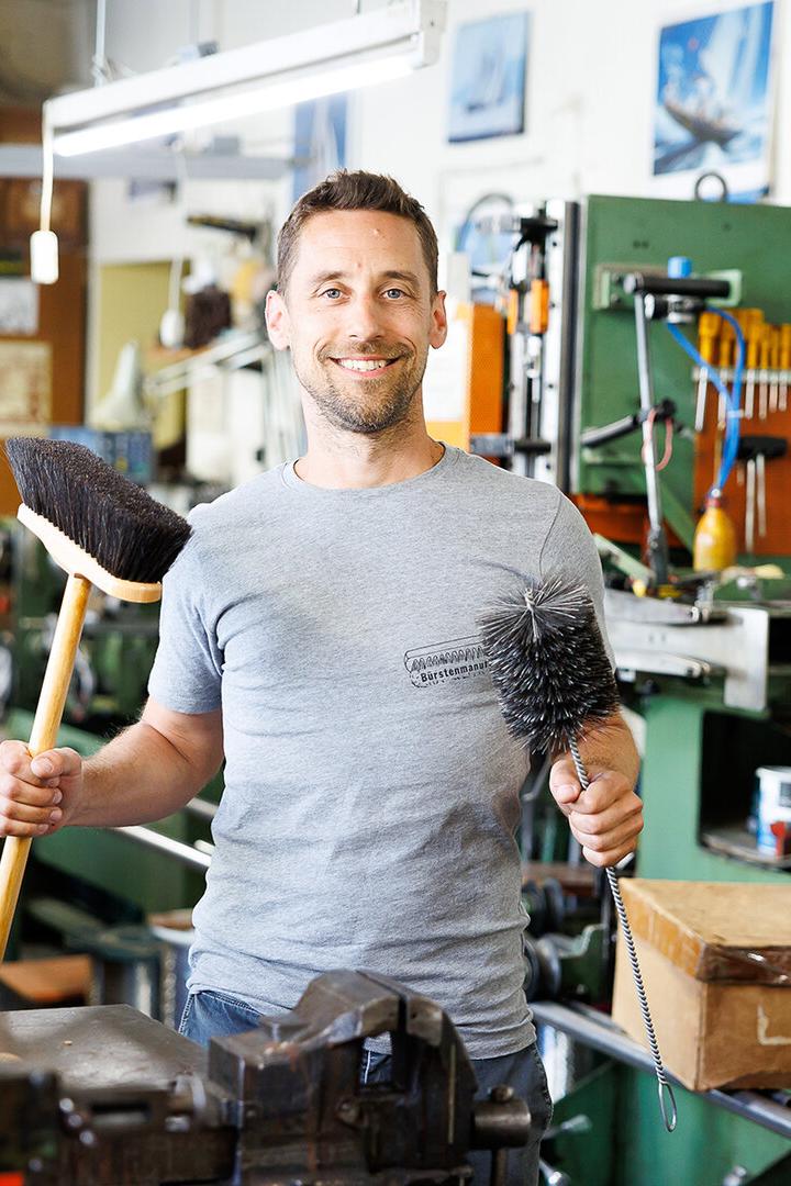 Ein Mann steht in einer Werkstatt und hält einen Besen und eine Bürste in den Händen, umgeben von Maschinen und Werkzeugen.