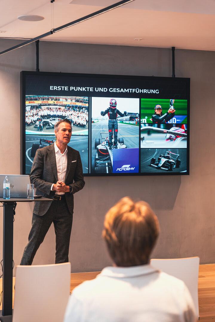Ein Mann im Anzug hält einen Vortrag vor einer Leinwand mit Motorsport-Bildern und der Überschrift „Erste Punkte und Gesamtführung“.