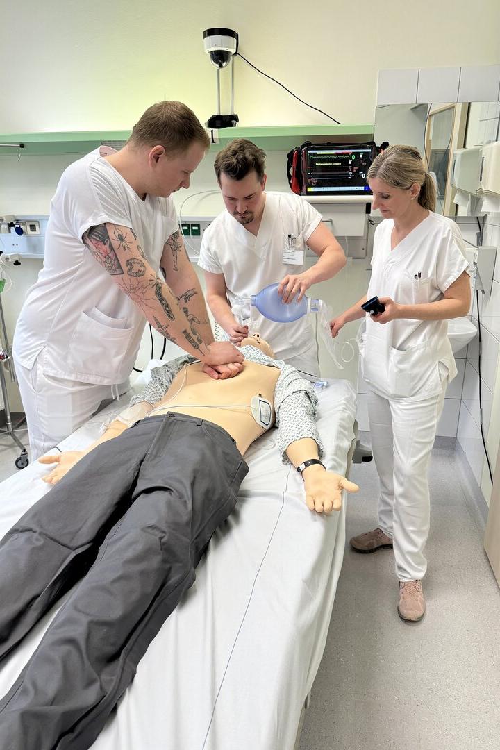 Drei Personen in weißer Kleidung üben an einer Reanimationspuppe Herz-Lungen-Wiederbelebung.