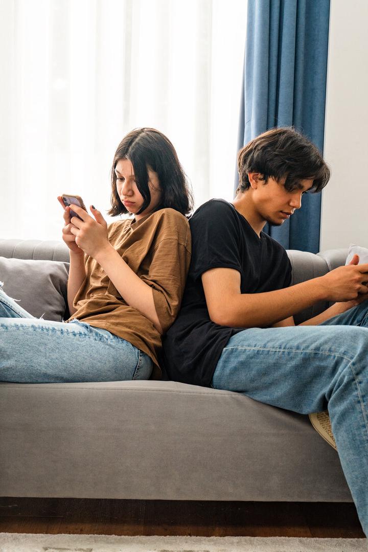 Zwei Jugendliche sitzen Rücken an Rücken auf einem Sofa und schauen konzentriert auf ihre Smartphones.