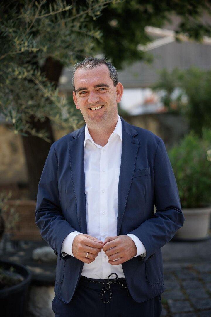 Ein Mann im blauen Anzug und weißem Hemd steht lächelnd im Freien vor grünen Pflanzen.