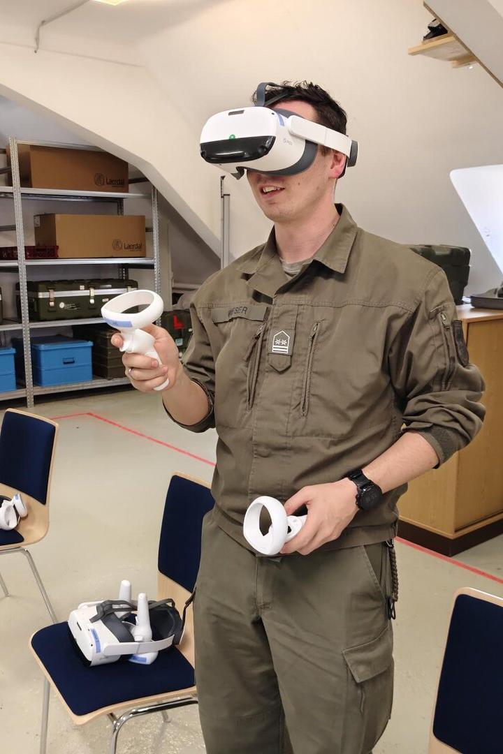 Eine Person in Uniform nutzt eine Virtual-Reality-Brille mit Controllern in einem Raum mit Stühlen und Regalen.