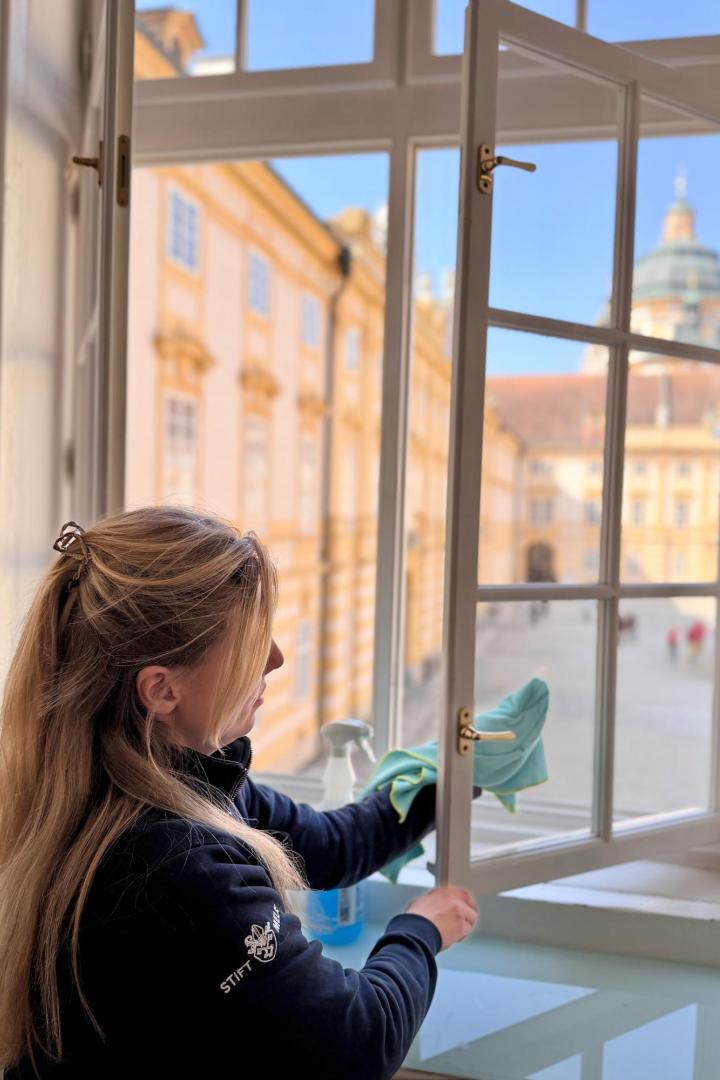 Frau reinigt ein geöffnetes Fenster im Stift Melk mit Blick auf den barocken Innenhof.
