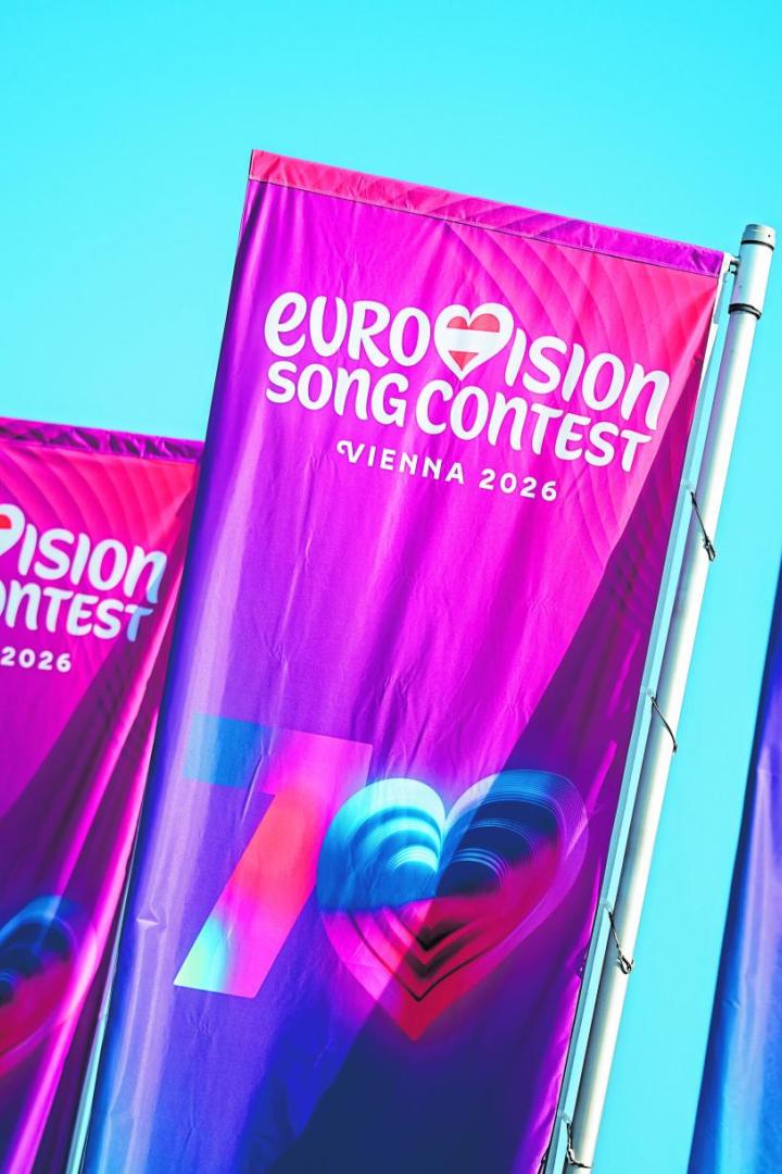 Mehrere bunte Fahnen mit der Aufschrift „Eurovision Song Contest Vienna 2026“ wehen vor blauem Himmel.