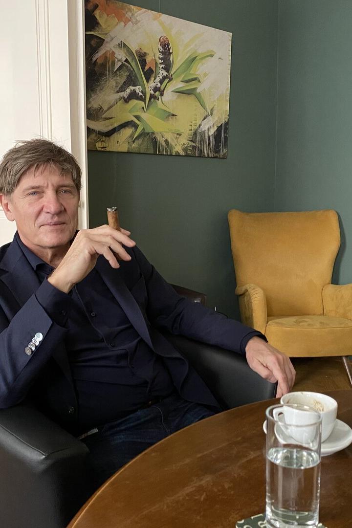 Ein Mann sitzt entspannt in einem Sessel, hält eine Zigarre, vor ihm stehen eine Tasse Kaffee und ein Glas Wasser auf dem Tisch.