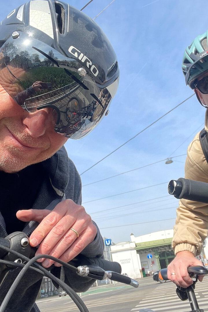 Zwei Männer mit Fahrradhelmen und Sonnenbrillen stehen mit ihren Fahrrädern an einer Straßenkreuzung bei sonnigem Wetter.