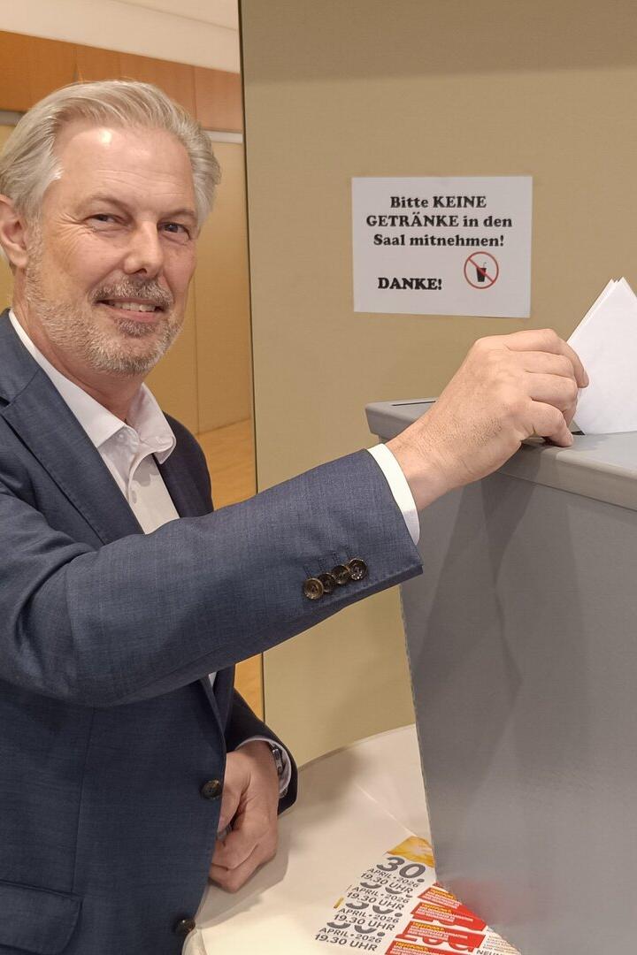 Ein Mann im Anzug wirft einen Wahlzettel in eine graue Wahlurne und lächelt dabei.