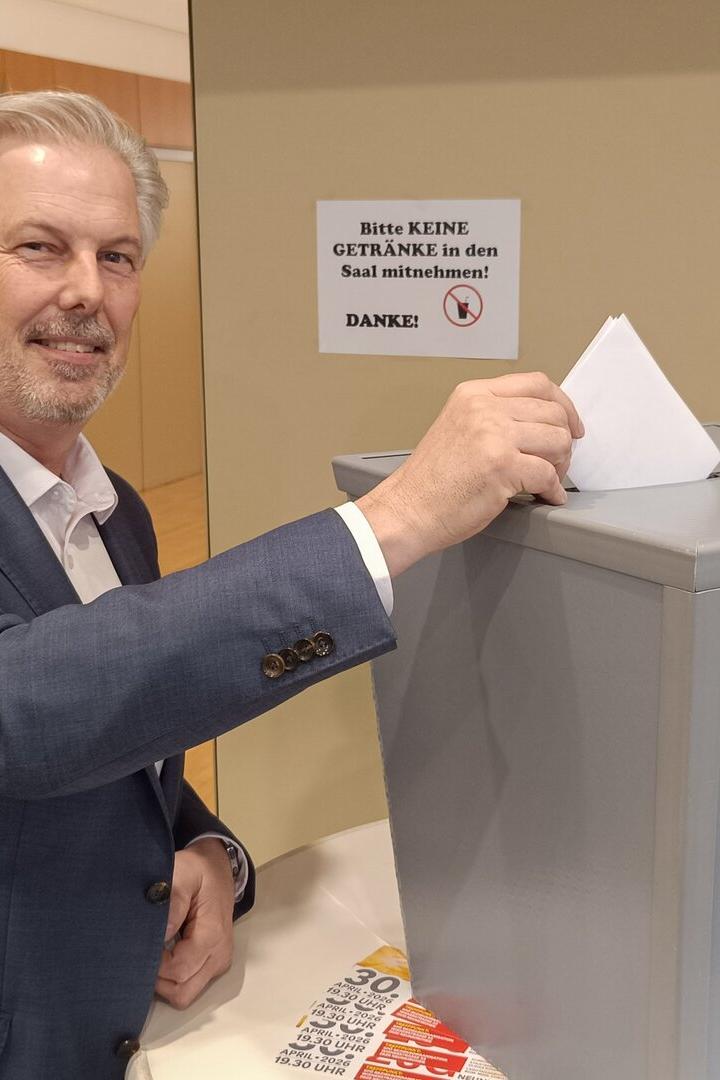 Ein Mann im Anzug wirft einen Wahlzettel in eine graue Wahlurne und lächelt dabei.