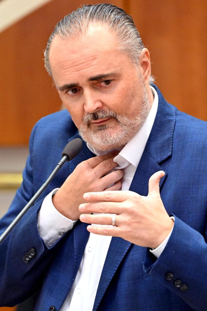 Ein Mann im blauen Anzug spricht gestikulierend an einem Mikrofon in einem Innenraum mit Holzverkleidung.