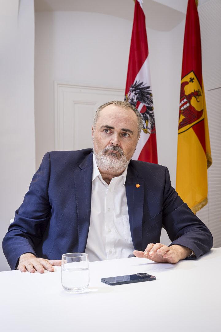 Ein Mann im Anzug sitzt an einem Tisch mit einem Glas Wasser und einem Smartphone, im Hintergrund zwei Fahnen.