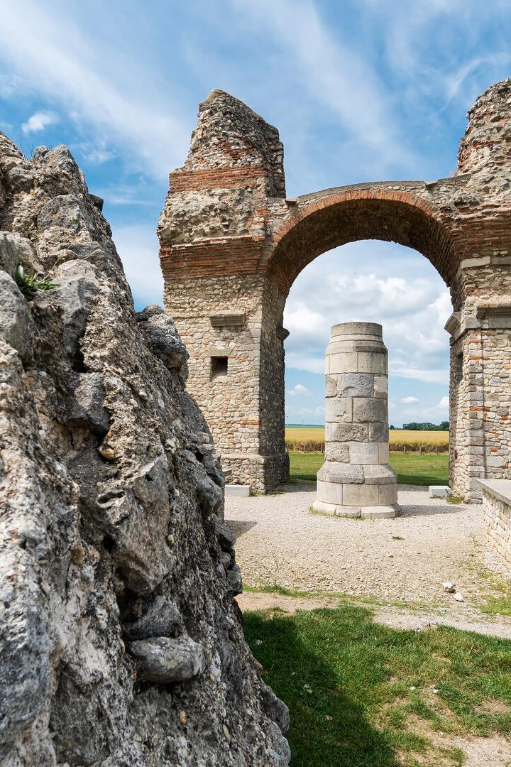 Eine alte Steinruine mit einem Bogen steht vor einem Feld unter blauem Himmel.