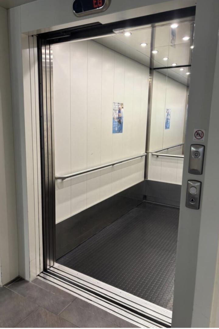 Ein offener Aufzug mit Spiegelwand, Haltegriff und schwarzem Boden ist zu sehen.