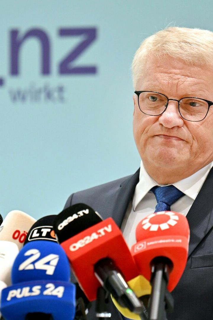 Ein Mann im Anzug steht vor mehreren Mikrofonen und spricht mit ernstem Gesichtsausdruck.