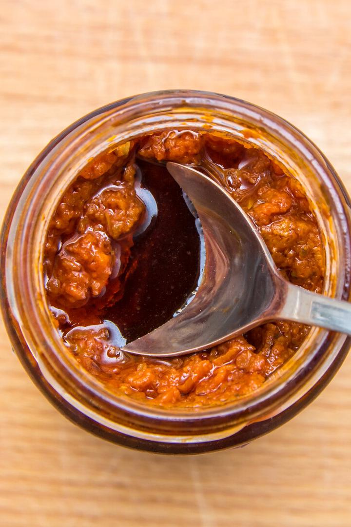 Ein Glas mit rotem Pesto und einem Löffel steht auf einem Holztisch.