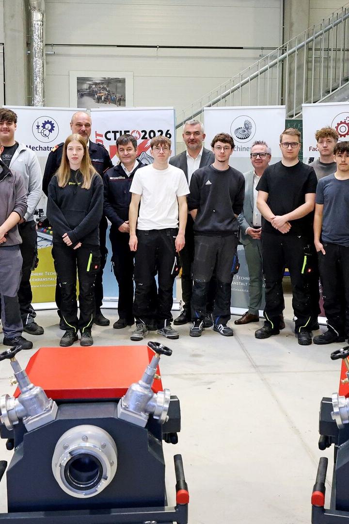 Eine Gruppe von jungen Erwachsenen und mehreren Erwachsenen steht in einer Werkhalle vor technischen Geräten und Informationsplakaten.