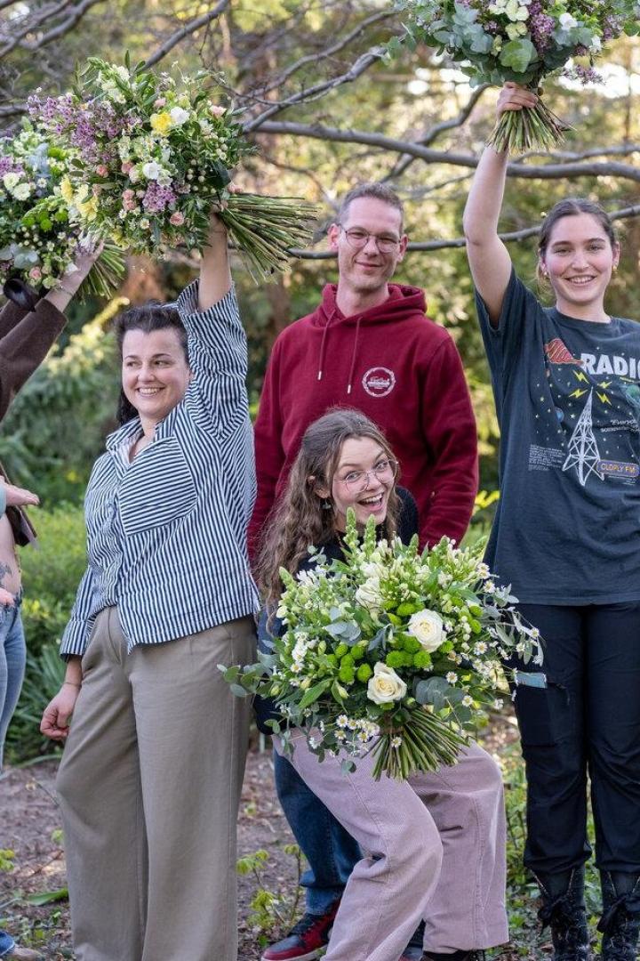 Sieben fröhliche Menschen posieren im Freien, einige halten große Blumensträuße in die Luft.