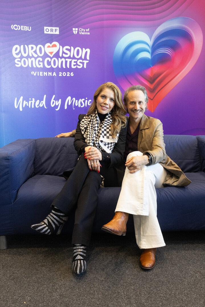 Zwei Personen sitzen vor einem Eurovision Song Contest 2026 Hintergrund auf einem Sofa.
