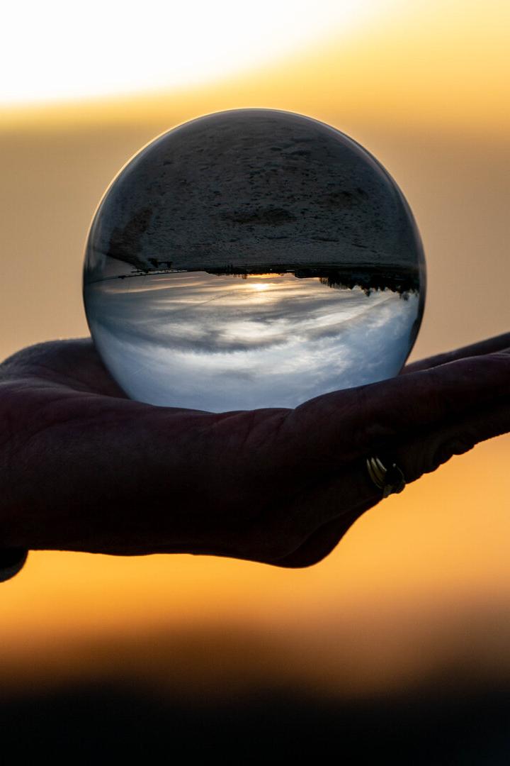 Eine Hand hält eine Glaskugel, in der eine Landschaft mit Sonnenuntergang auf dem Kopf gespiegelt wird.