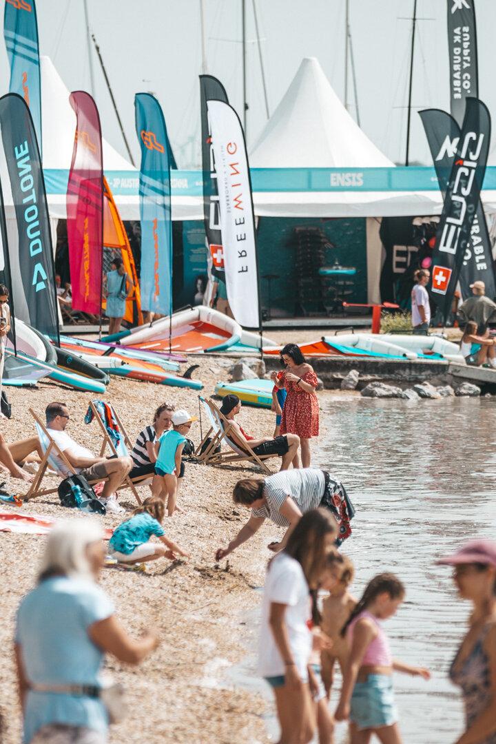 Viele Menschen entspannen am Strand, spielen im Wasser und bunte Fahnen sowie Zelte stehen im Hintergrund.